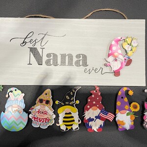May include: A wooden sign with the text "Best Nana ever" is surrounded by colorful gnome cutouts. The gnomes have various designs, including floral, patriotic, and seasonal themes. The sign has a jute rope for hanging.