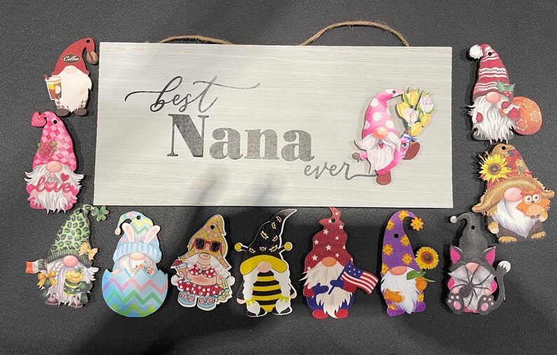 May include: A wooden sign with the text "Best Nana ever" is surrounded by colorful gnome cutouts. The gnomes have various designs, including floral, patriotic, and seasonal themes. The sign has a jute rope for hanging.