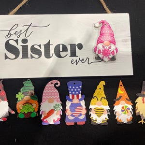May include: A white wooden sign with the text "best Sister ever" in black script and a pink gnome. Below the sign are eight gnome cutouts with various designs, including hearts, shamrocks, and patriotic themes.