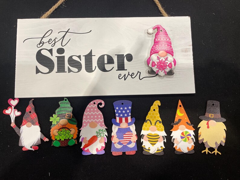 May include: A white wooden sign with the text "best Sister ever" in black script and a pink gnome. Below the sign are eight gnome cutouts with various designs, including hearts, shamrocks, and patriotic themes.