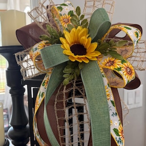 May include: A decorative bow featuring a large yellow sunflower with a dark brown center, surrounded by green foliage. The bow is crafted from layers of solid brown ribbon, natural burlap mesh, and a white ribbon printed with sunflowers.