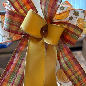 May include: A decorative bow crafted from multiple ribbons, featuring a mix of solid mustard yellow fabric and patterned ribbons with orange, green, and yellow plaid designs, alongside a white ribbon printed with orange pumpkins and green vines.