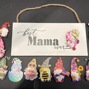 May include: A white wooden sign with the words "Best Mama ever" and a jute rope hanger. The sign is surrounded by various gnome cutouts with colorful hats and outfits, including a gnome holding an American flag.