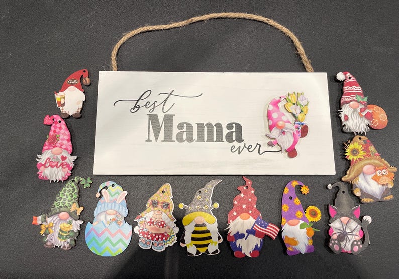 May include: A white wooden sign with the words "Best Mama ever" and a jute rope hanger. The sign is surrounded by various gnome cutouts with colorful hats and outfits, including a gnome holding an American flag.