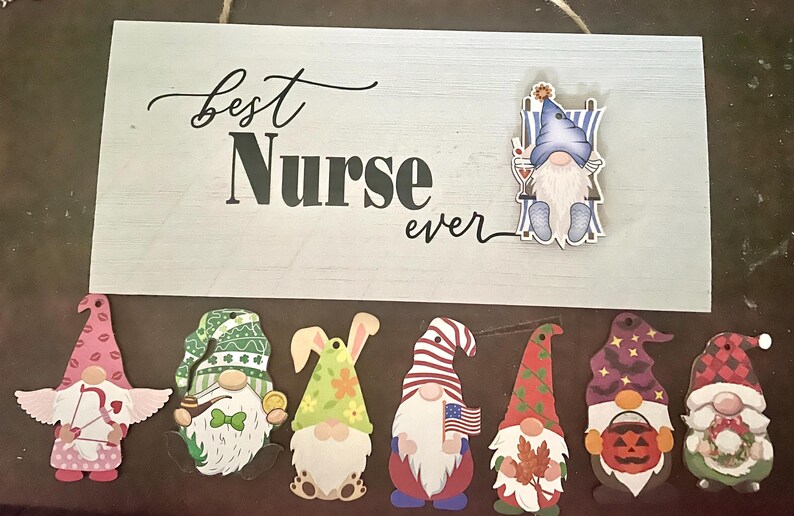 May include: A wooden sign with the words "Best Nurse ever" and a collection of gnome figurines. The gnomes are in various designs, including a nurse gnome, a cupid gnome, and gnomes with patriotic and seasonal themes.