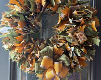 May include: A circular wreath constructed from numerous fabric strips in shades of mustard yellow, olive green, and white, featuring patterns of sunflowers, polka dots, and stripes, finished with a large, multi-layered fabric bow at the bottom.