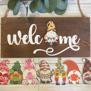 May include: A wooden welcome sign with the word "welcome" in white lettering.  The sign is decorated with eight gnome ornaments, each with a different theme, including a pumpkin, a sunflower, a shamrock, an Easter egg, and a USA flag.