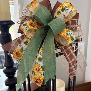 May include: A decorative multi-layered bow featuring a combination of solid brown ribbon, a sunflower-patterned ribbon with yellow petals and green leaves, a solid sage green ribbon, and a natural tan jute mesh ribbon tied together in a cluster.