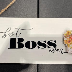 May include: White wooden sign with the words "best Boss ever" in black script and block letters. A decorative image of flowers and bees is on the right side. A brown rope is attached for hanging.