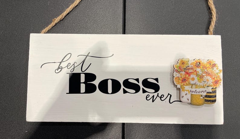 May include: White wooden sign with the words "best Boss ever" in black script and block letters. A decorative image of flowers and bees is on the right side. A brown rope is attached for hanging.