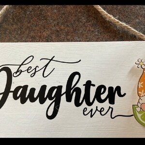 May include: A white wooden sign with the words "best Daughter ever" in black script. A gnome with an orange hat and a lime slice is on the right side. The sign is hanging by a natural rope.
