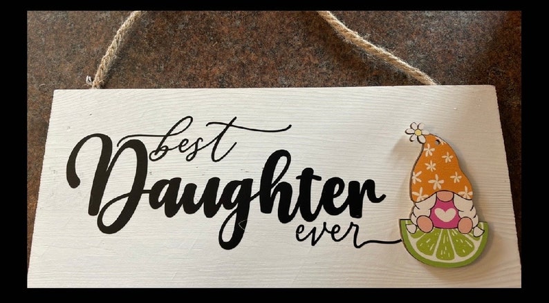 May include: A white wooden sign with the words "best Daughter ever" in black script. A gnome with an orange hat and a lime slice is on the right side. The sign is hanging by a natural rope.