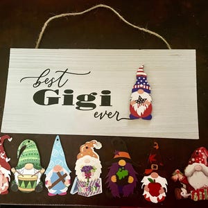 May include: A white wooden sign with the words "best Gigi ever" in black script and block letters. The sign is decorated with a gnome wearing a patriotic hat and several other gnome ornaments in various colors and designs.