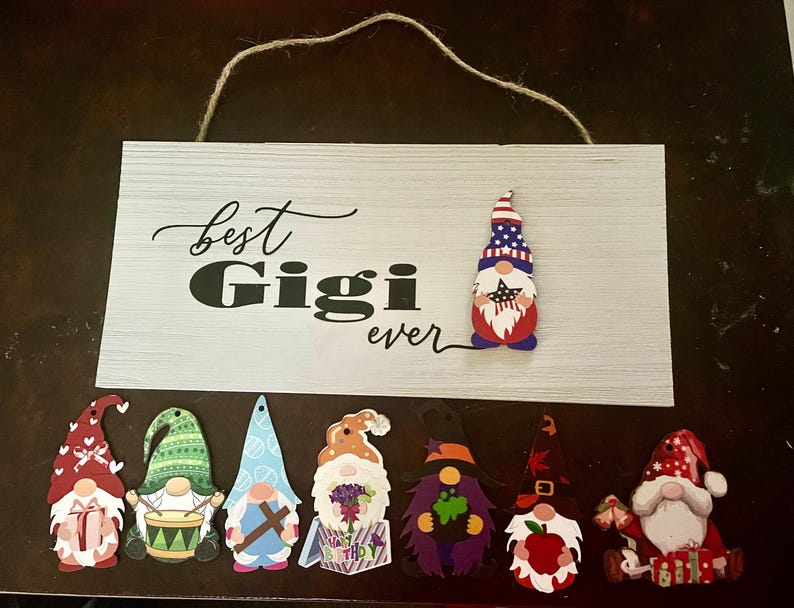 May include: A white wooden sign with the words "best Gigi ever" in black script and block letters. The sign is decorated with a gnome wearing a patriotic hat and several other gnome ornaments in various colors and designs.