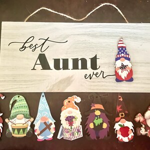 May include: A wooden sign with the text "Best Aunt ever" and several gnome cutouts. The sign has a light gray wood grain pattern. The gnomes are in various colors and designs, including a patriotic gnome.