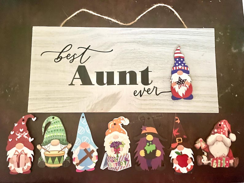 May include: A wooden sign with the text "Best Aunt ever" and several gnome cutouts. The sign has a light gray wood grain pattern. The gnomes are in various colors and designs, including a patriotic gnome.