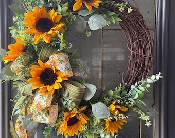 May include: A circular wreath constructed from brown twigs, adorned with bright yellow sunflowers, green eucalyptus leaves, and small white floral sprigs. A decorative ribbon featuring a sunflower pattern and green stripes is tied to the side.