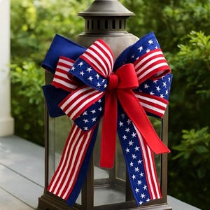 May include: A decorative lantern adorned with a patriotic bow. The bow features red and white striped ribbon, blue ribbon with white stars, and a solid red center. The lantern is dark brown and sits on a wooden surface.