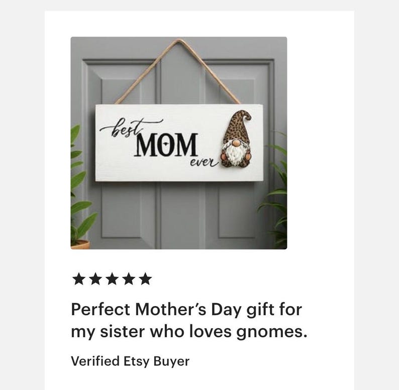 May include: A white wooden sign with the words "best MOM ever" in black script and block letters. A gnome with a brown and tan hat and white beard is on the right. The sign hangs from a natural rope.