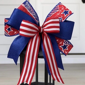 May include: A patriotic bow with red, white, and blue ribbons. The bow features a striped center and star-patterned accents, resting atop a black lantern. The bow's design incorporates the colors of the American flag.