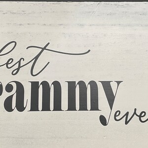 May include: A white wooden sign with the words "best Grammy ever" in black script. A green gnome with a rainbow and shamrock is attached to the right side of the sign. The sign has a distressed, rustic look.