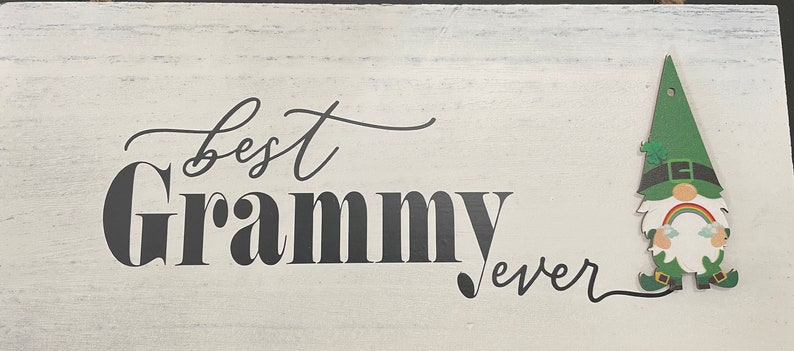 May include: A white wooden sign with the words "best Grammy ever" in black script. A green gnome with a rainbow and shamrock is attached to the right side of the sign. The sign has a distressed, rustic look.
