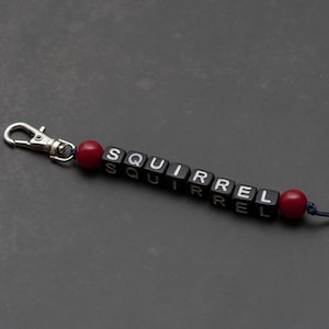 Könnte beinhalten: Ein Schlüsselanhänger mit dem Wort "SQUIRREL" in schwarzen Buchstabenperlen. Der Schlüsselanhänger hat einen silbernen Verschluss und ist mit roten Perlen akzentuiert. Der Hintergrund ist ein dunkles Grau.