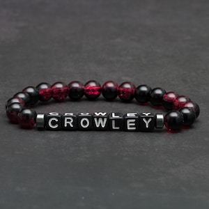 May include: A beaded bracelet with the name "CROWLEY" spelled out in white letters on black square beads. The bracelet features alternating black and red-toned beads, set against a dark background.