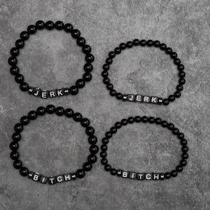 Puede incluir: Cuatro pulseras de cuentas negras con cuentas blancas con las palabras "JERK" y "BITCH". Las pulseras están dispuestas sobre una superficie gris.
