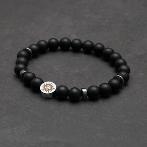 Op de afbeelding: Zwart kralenarmband met zilveren accent. De armband heeft matte zwarte kralen en zilverkleurige afstandhouders. Het zilveren element heeft een zonmotief. Een stijlvol accessoire.