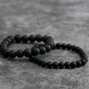 May include: Two black beaded bracelets. One bracelet has larger beads, the other smaller. The bracelets are made of a matte material and are displayed on a grey surface. These bracelets are a stylish accessory.
