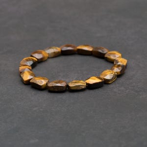 May include: A stretch bracelet composed of faceted, irregular tiger's eye beads. The beads display a natural chatoyant lustre with alternating bands of golden-brown and dark brown tones, arranged in a circular loop on a dark grey surface.