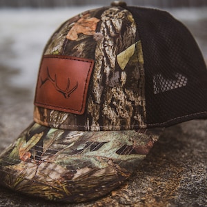 Puede incluir: Una gorra de camionero de camuflaje con un parche de cuero marrón que presenta un diseño de asta de alce. La gorra tiene una parte posterior de malla negra.