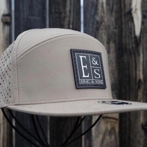 May include: A tan baseball cap with a flat brim and a perforated side panel. The cap features a black square patch with the text "E & S ERIC & SONS" in white.