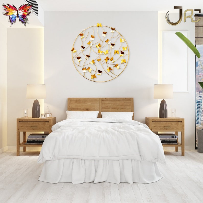 May include: A white bedroom with a gold metal wall decor featuring a circle with butterflies. The decor is hanging on a white wall above a wooden bed with white bedding. There are two wooden nightstands with lamps on either side of the bed.