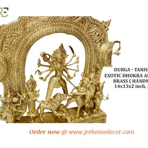 May include: A gold-colored metal sculpture of the Hindu goddess Durga, standing on a lion, with multiple arms and holding weapons. The sculpture is framed by a decorative archway. The text "DURGA - TANISHQA EXOTIC DHOKRA ARTIFACT BRASS (HANDMADE) 14x13x2 inch, 3Kg" is visible below the sculpture.