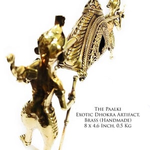 May include: A gold-colored metal sculpture of a palanquin, a traditional Indian ceremonial litter, carried by two figures. The palanquin is decorated with intricate details and measures 8 inches by 4.6 inches. The sculpture is made of brass and weighs 0.5 kg. "THE PAALKI EXOTIC DHOKRA ARTIFACT, BRASS (HANDMADE) 8 X 4.6 INCH, 0.5 KG" is written below the sculpture.