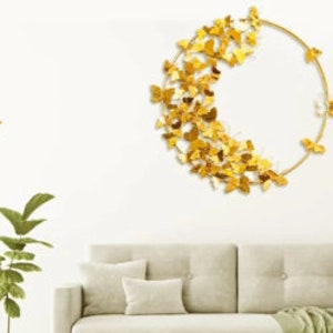 May include: Gold metal wall decor featuring a circle with a cluster of butterflies. The butterflies are arranged in a crescent shape on the left side of the circle. The decor is hanging on a white wall above a beige sofa.