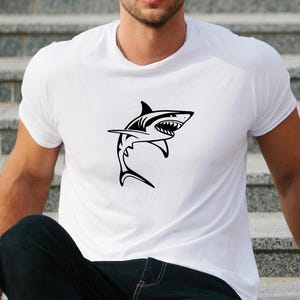 Puede incluir: Una camiseta blanca con un gr&aacute;fico de silueta de tibur&oacute;n negro.