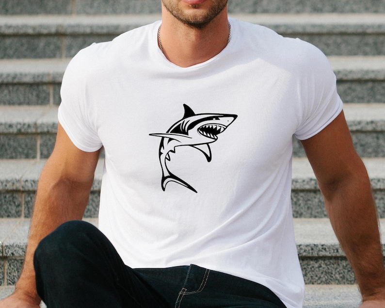 Puede incluir: Una camiseta blanca con un gr&aacute;fico de silueta de tibur&oacute;n negro.