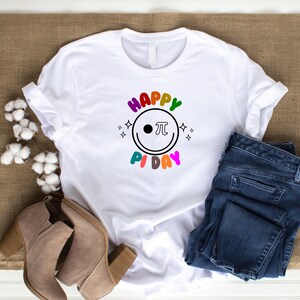 K&ouml;nnte beinhalten: Wei&szlig;es T-Shirt mit einer regenbogenfarbenen Grafik, auf der "Happy Pi Day" mit einem Smiley und einem Pi-Symbol steht.