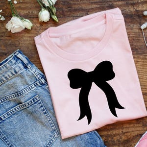 May include: A pale pink t-shirt with a black bow design. The image includes blue denim jeans, a pink trainer, white flowers, and earbuds, all arranged on a wooden surface.