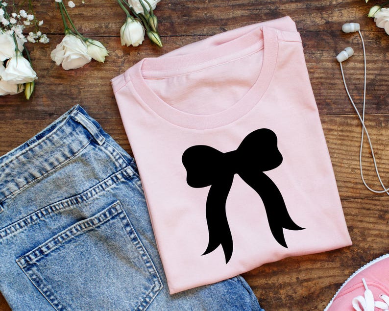 May include: A pale pink t-shirt with a black bow design. The image includes blue denim jeans, a pink trainer, white flowers, and earbuds, all arranged on a wooden surface.
