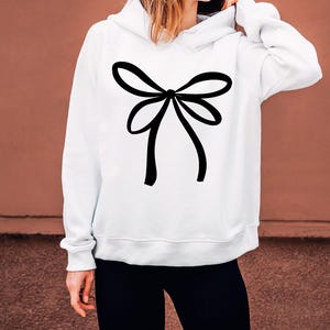 May include: White hoodie featuring a large black bow design on the front. The bow is centrally placed and has a simple, elegant style. The hoodie has a drawstring hood and long sleeves. The background is a solid brown colour.