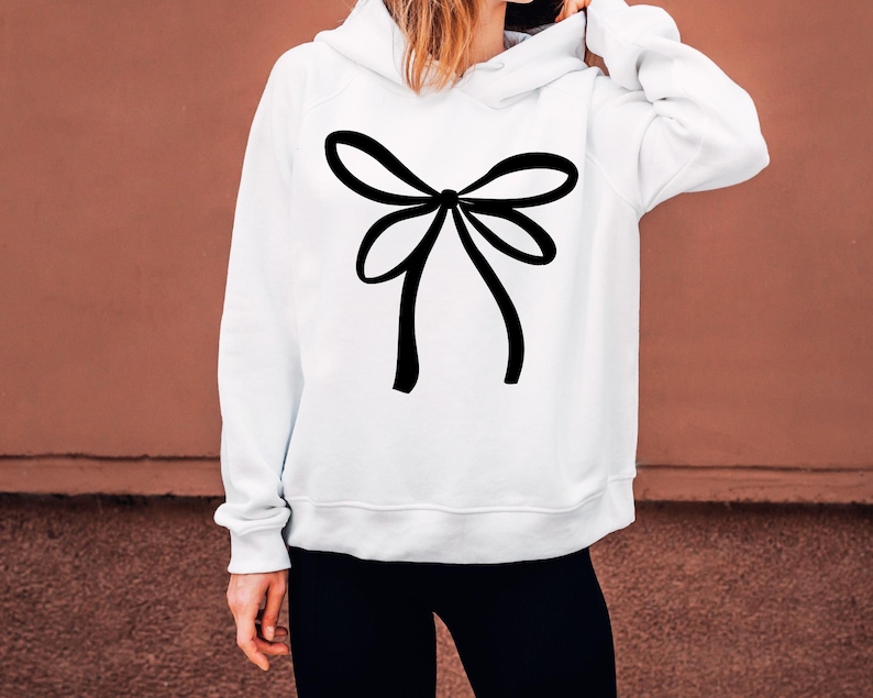 May include: White hoodie featuring a large black bow design on the front. The bow is centrally placed and has a simple, elegant style. The hoodie has a drawstring hood and long sleeves. The background is a solid brown colour.