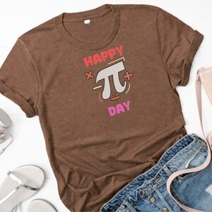 K&ouml;nnte beinhalten: Ein braunes T-Shirt mit dem Text "Happy Pi Day" in Rosa, Rot und Orange. Der Text ist von mathematischen Symbolen umgeben.