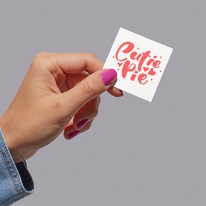 K&ouml;nnte beinhalten: Eine Hand h&auml;lt eine wei&szlig;e Karte mit dem Text "cutie pie" in rosa kursiver Schrift. Die Karte ist mit kleinen rosa Herzen verziert.
