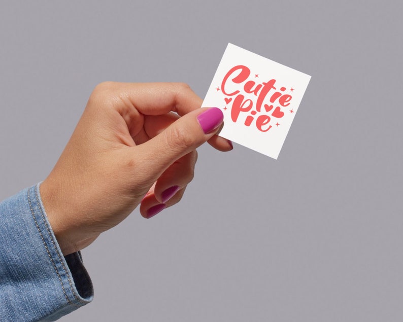 K&ouml;nnte beinhalten: Eine Hand h&auml;lt eine wei&szlig;e Karte mit dem Text "cutie pie" in rosa kursiver Schrift. Die Karte ist mit kleinen rosa Herzen verziert.