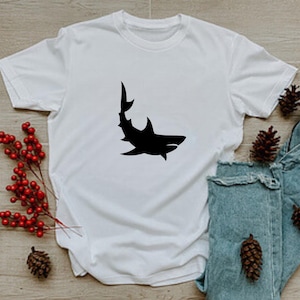 Puede incluir: Camiseta blanca con una silueta negra de un tibur&oacute;n en la parte delantera.