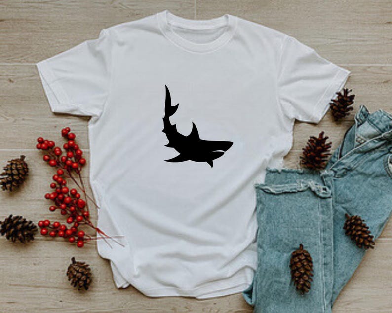 Puede incluir: Camiseta blanca con una silueta negra de un tibur&oacute;n en la parte delantera.